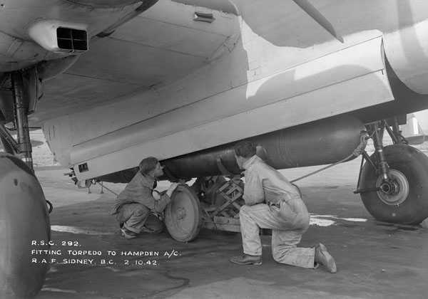 Black and white photograph. Underneath an airplane, both large wheels visible. A compartment is open, and two men crouch down to load a large bomb (6-8 feet long) into the plan. It is mounted on a jack.
